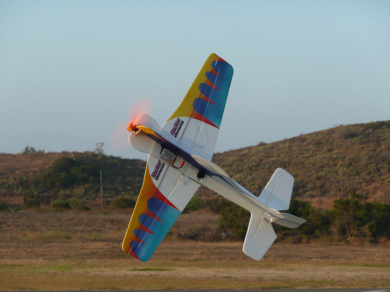 Colorful model airplane performing a maneuver on a grassy field with hills in the background.