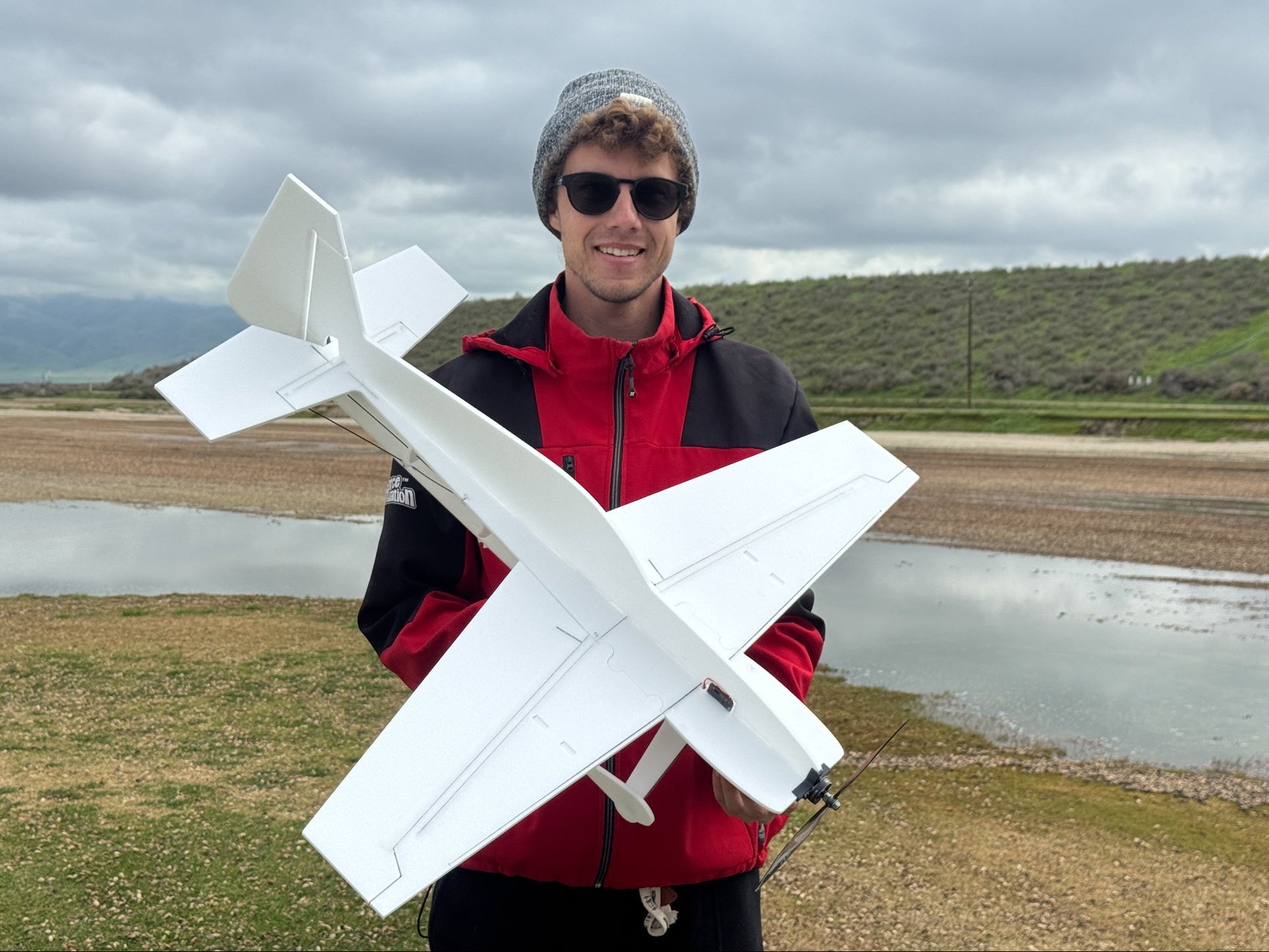 Matt Stringer holding a large white model airplane outdoors with a cloudy sky and grassy field in the background.