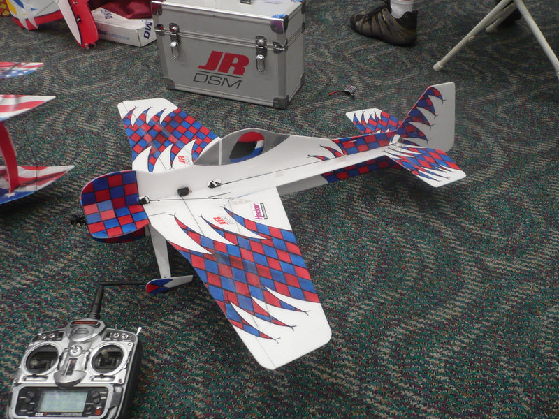 Model airplane with red and blue checkered pattern on a carpeted floor, accompanied by a remote control.