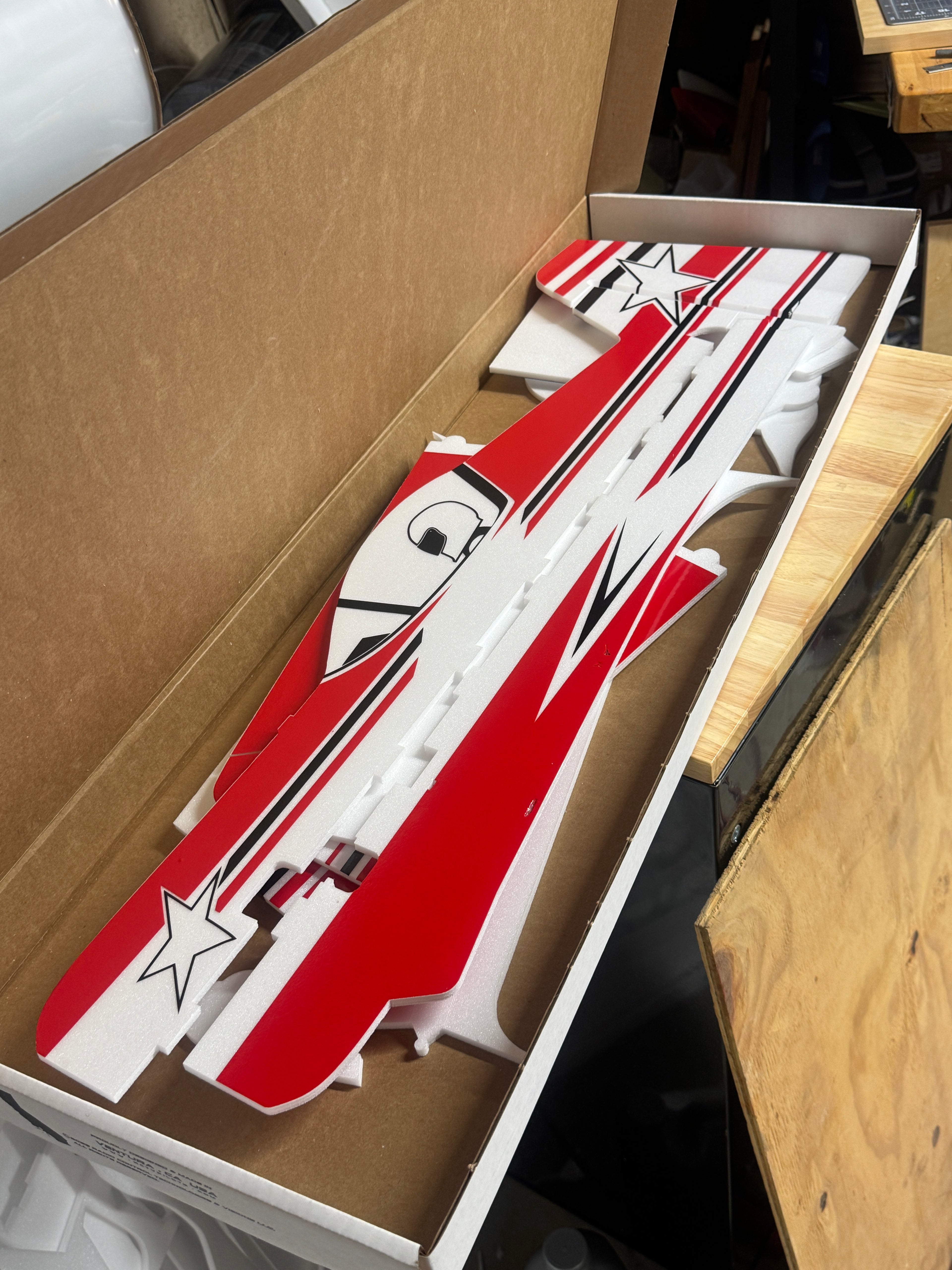 Red and white model airplane kit inside a cardboard box on a wooden surface.