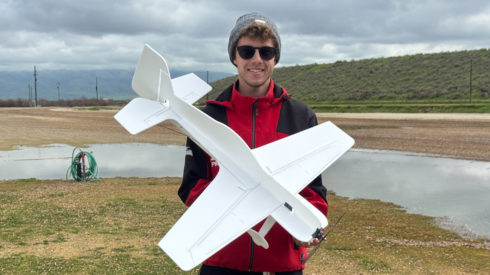 Matt Stringer holding a large white model airplane outdoors with a cloudy sky and landscape in the background.